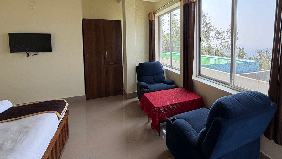 A guest bedroom featuring blue armchairs, a small table with a red cloth, and large windows looking outside | Kollanoor Guest House, Coorg
