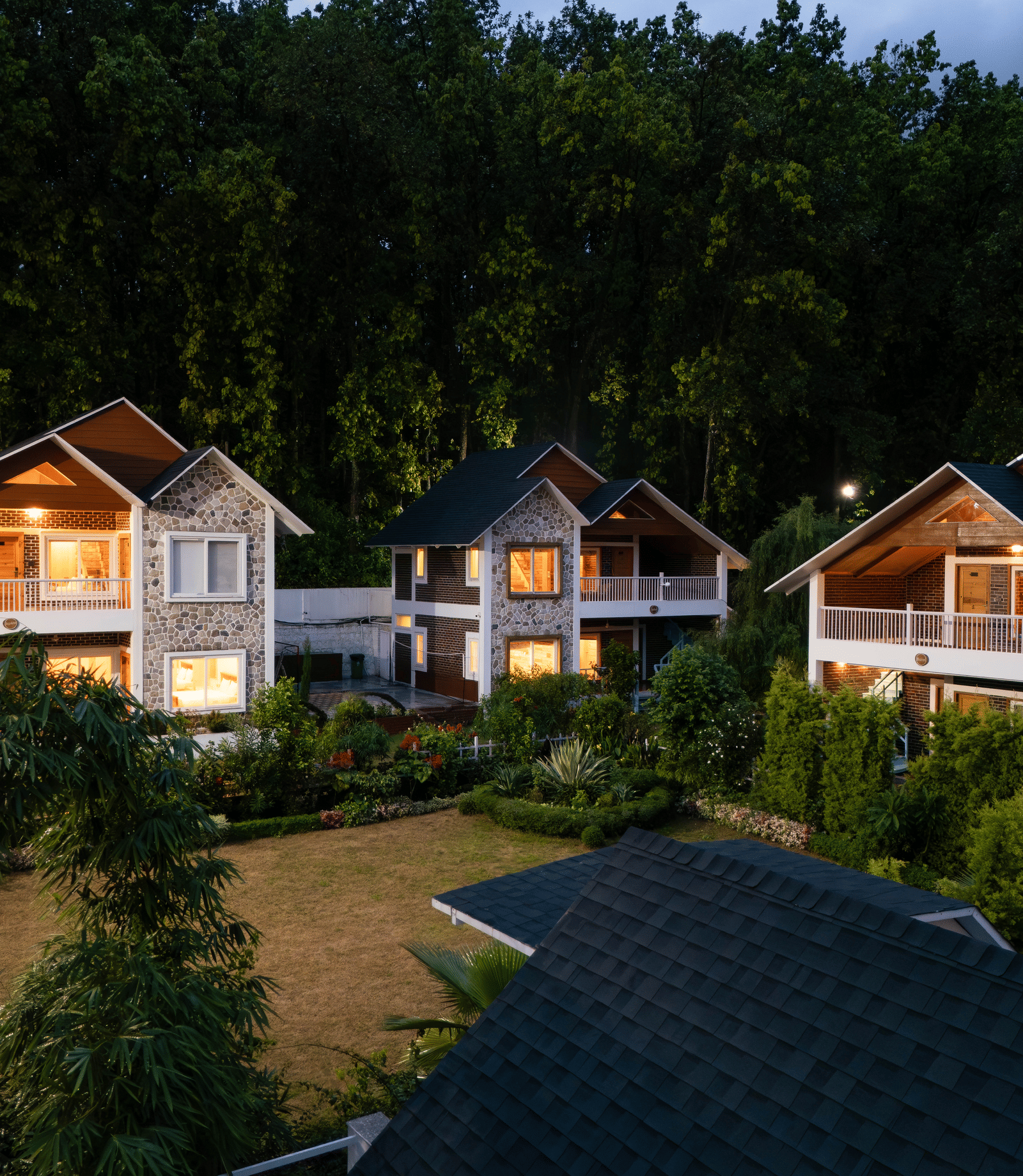 An evening exterior shot of a row of modern, stone-clad villas nestled among dark green trees - Megha Resort & Villas, Dehradun