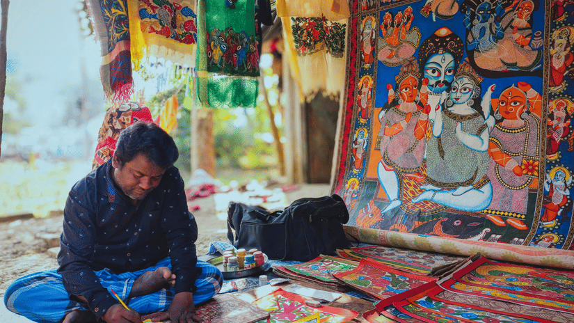 A person sits on the ground and paints a detailed piece of art. Various cloths and finished paintings hang in the background.