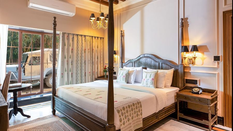 Elegantly designed hotel room featuring a four-poster wooden bed with floral cushions and matching bed runner, classic wall panelling, warm lighting from wall sconces and chandelier, a ceiling fan, and a sliding glass door revealing a parked jeep outside -