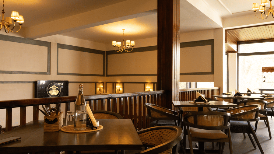 A dining room seen from behind a wooden railing, featuring circular tables, cushioned chairs, and framed artwork on the walls under soft interior lighting.| The Heritage Club, Tripura, Castle, Shillong