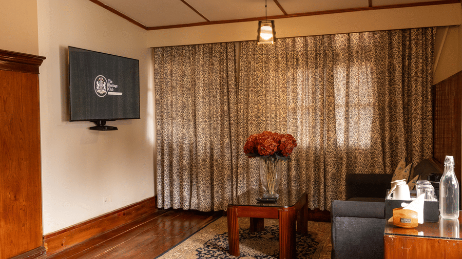 A room at The Heritage Club, Tripura, Castle, Shillong, showcasing a flower vase placed on a table, sofas, a television, and a large window covered with curtains.