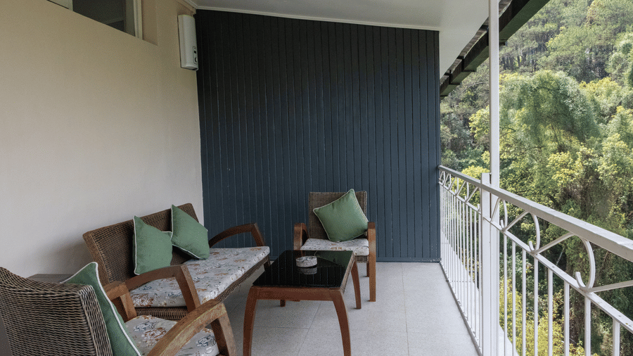 A balcony in a room at The Heritage Club, Tripura, Castle, Shillong, featuring pillows placed on chairs with cushions, a table, and trees in the front.