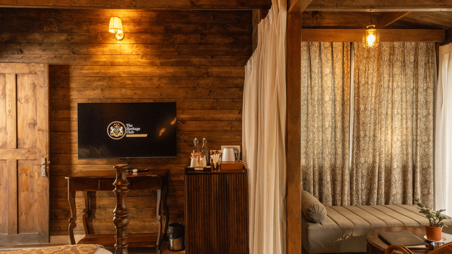A room at The Heritage Club, Tripura, Castle, Shillong, featuring a television displaying the hotel name, a wall dividing the room into two, on one side exists a bed, a pillow, the other side contains a chair and a table.