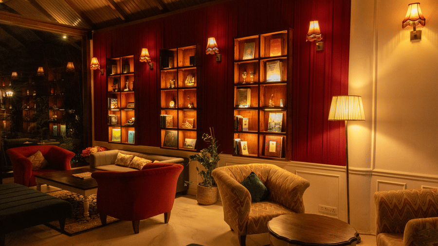 A cozy lounge area with warm, ambient lighting, plush armchairs, and a large bookshelf-style display set against a dark red accent wall. |  The Heritage Club, Tripura, Castle, Shillong