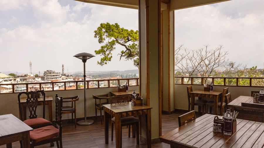 An outdoor terrace dining space with wooden tables and chairs, offering a view of the surrounding cityscape under an overcast sky. |  The Heritage Club, Tripura, Castle, Shillong