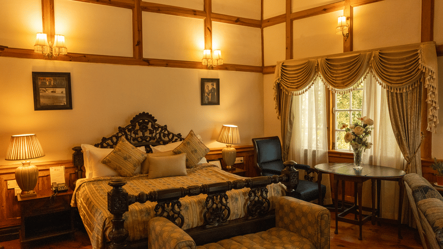 A room at The Heritage Club, Tripura, Castle, Shillong, showcasing a large bed, pillows, spreads, lamps, windows, and tables.