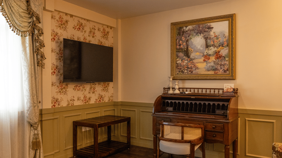 A room at The Heritage Club, Tripura, Castle, Shillong, showcasing a wall-mounted television, wall art, tables, a chair, and a window.