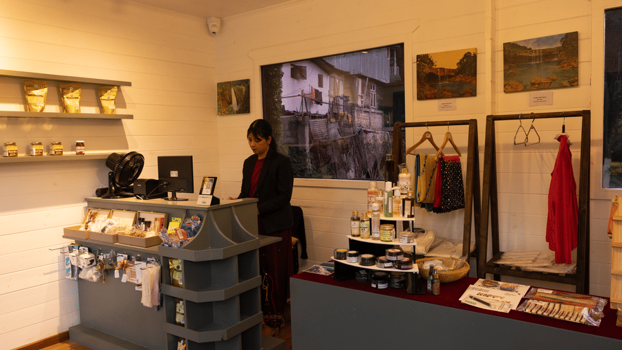 An illuminated Souvenir shop with a woman standing near the table, clothes, and other small items placed on a shelf. | The Heritage Club, Tripura, Castle, Shillong