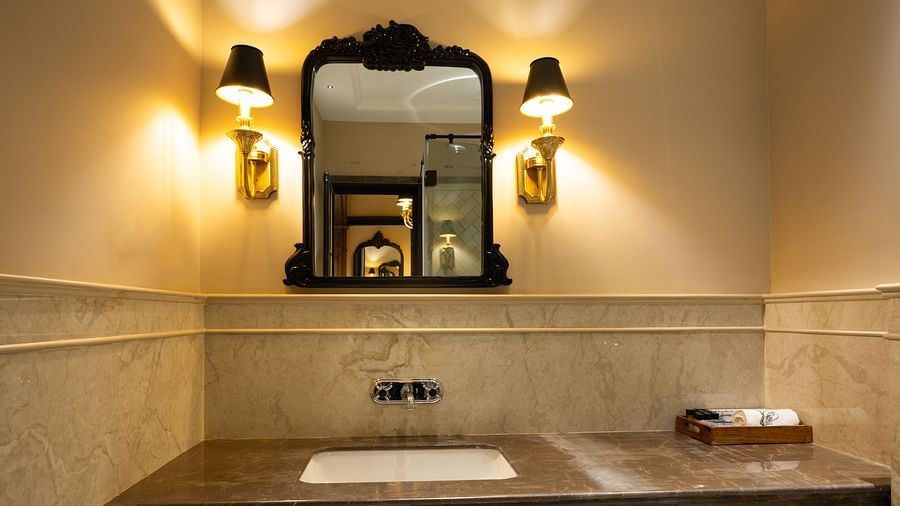 Bathroom vanity with a dark framed mirror, wall-mounted lamps on both sides, and a countertop with a built-in sink and toiletry tray - Panna Vilas Palace
