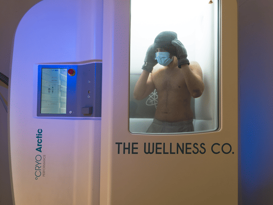 A man wearing a mask stands inside a cryotherapy chamber labeled with a lit digital control panel on the left.