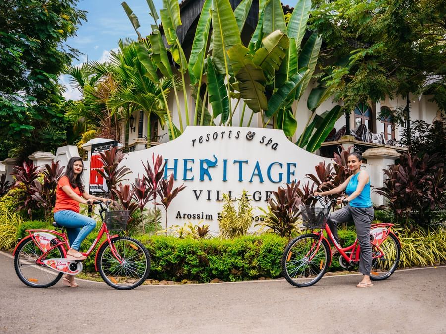 Guests riding bicycles past the main entrance signage of Heritage Village Resorts & Spa, Goa.