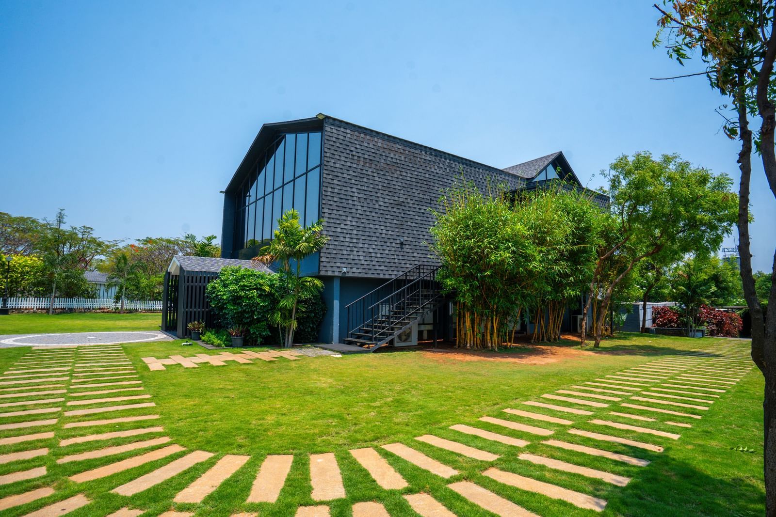 Facade of Love Letter at Darza Resort with large windows is nestled amongst bright green lawns and trees under a clear, sunny sky, with stone pathways leading across the grass.