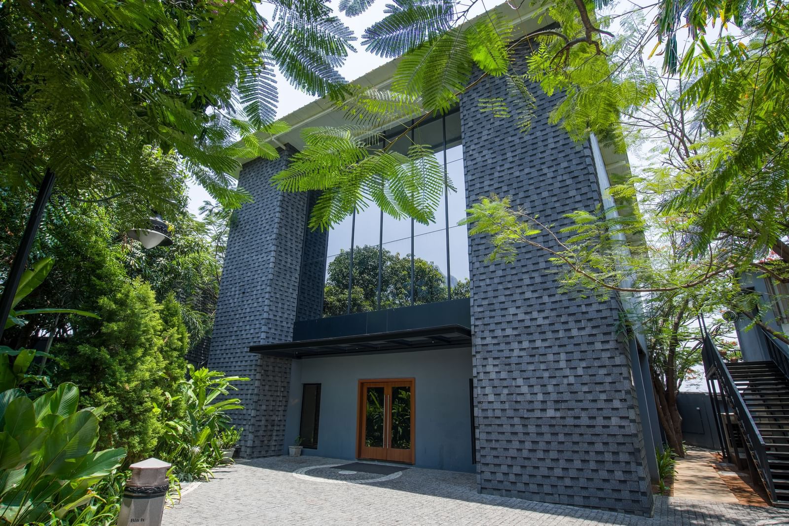 Facade of Royal Affair at Darza Resort with textured grey walls and large glass windows is partially obscured by lush green foliage under a bright sky.
