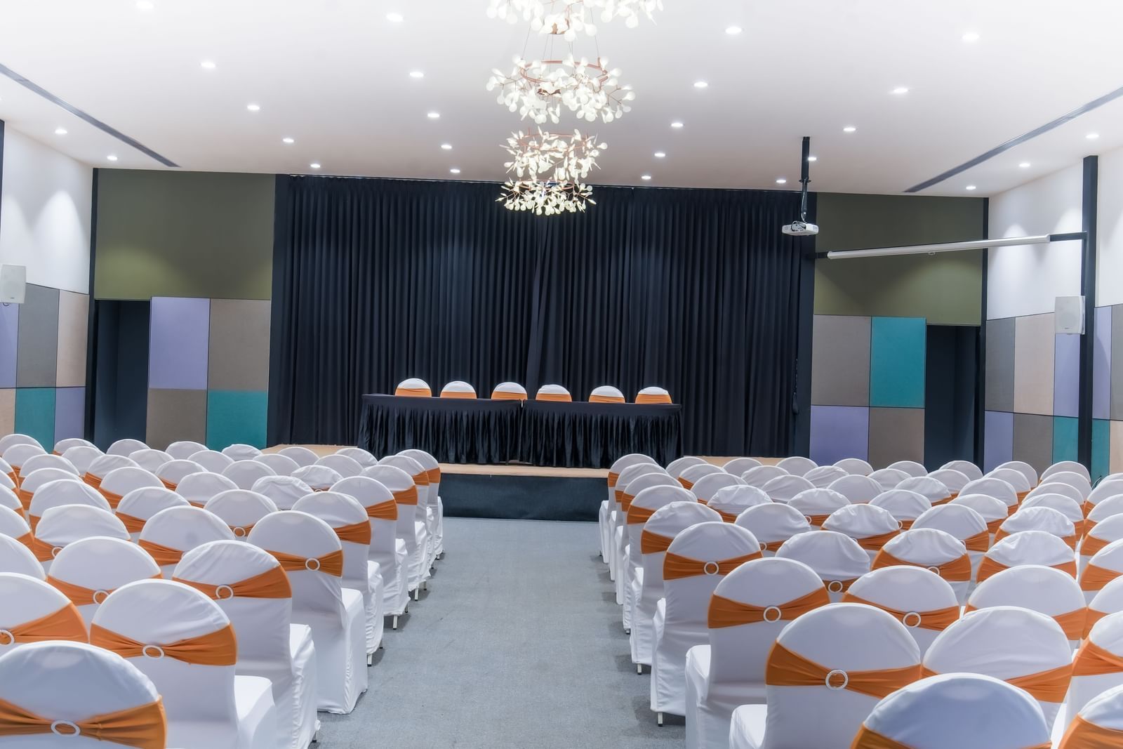 Royal Affair at Darza Resort features numerous white chairs indoor with orange sashes facing a stage with a draped backdrop and several chairs.