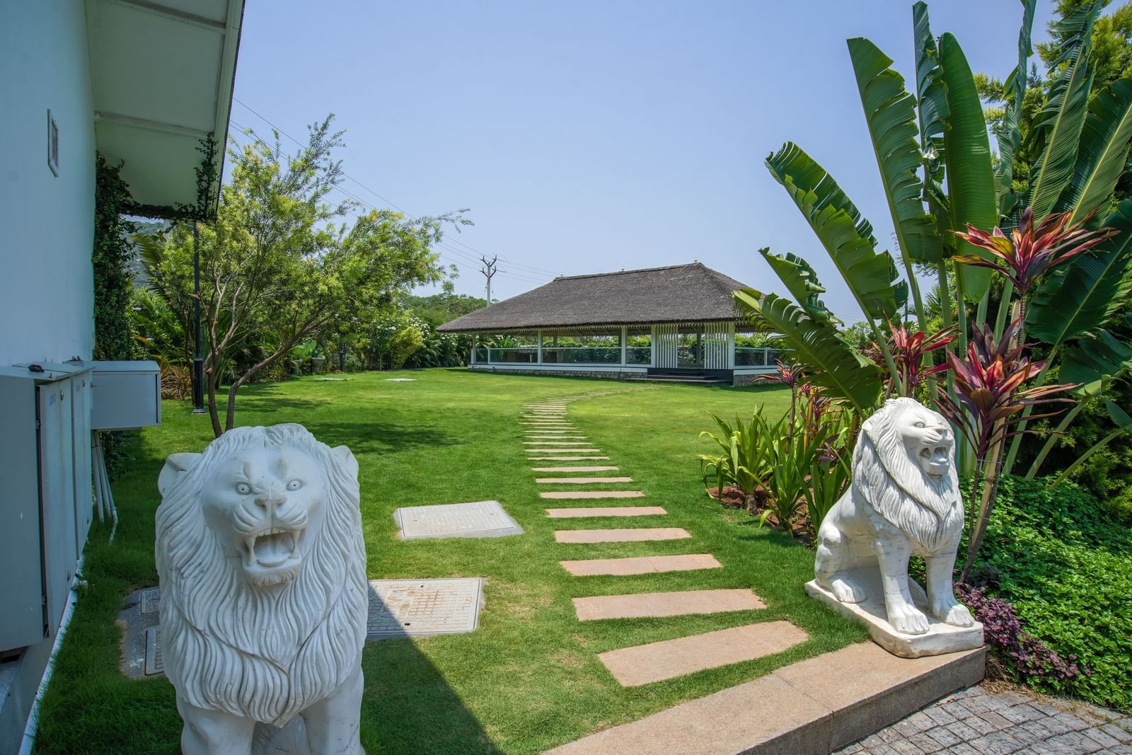 Slice of Heaven at Darza Resort featuring a stone pathway, flanked by two white lion statues, leads across a green lawn towards an open-sided building amidst lush tropical foliage under a bright sky.