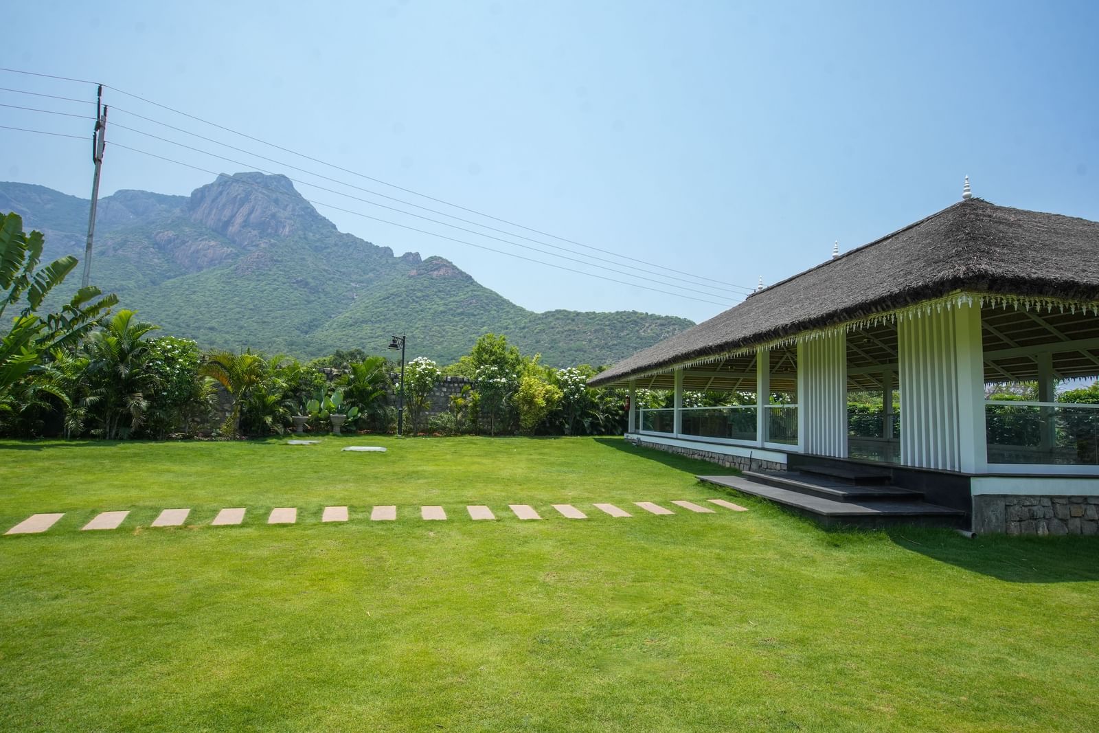 Slice of Heaven at Darza Resort featuring a bright green lawn with a stone pathway leads to a white building with a thatched roof, set against a backdrop of lush greenery and mountains.