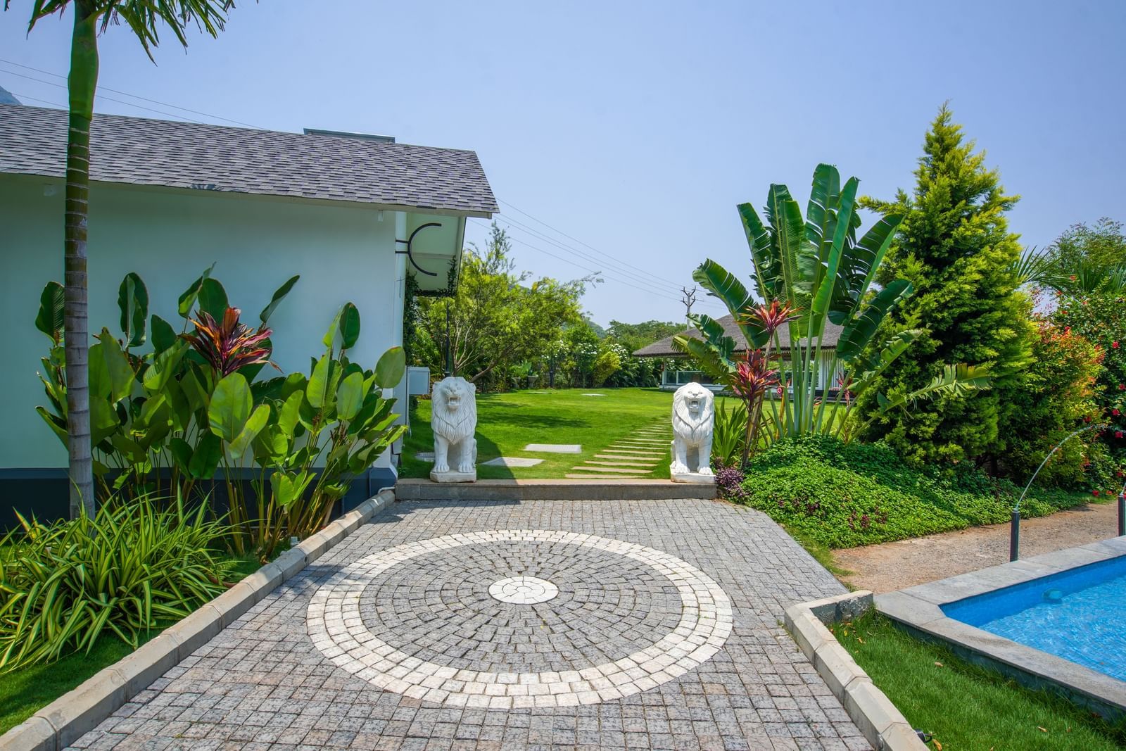 Slice of Heaven at Darza Resort featuring stone pathway leading past two white elephant statues towards a green lawn and lush tropical plants under a bright sky, with a glimpse of a blue pool.