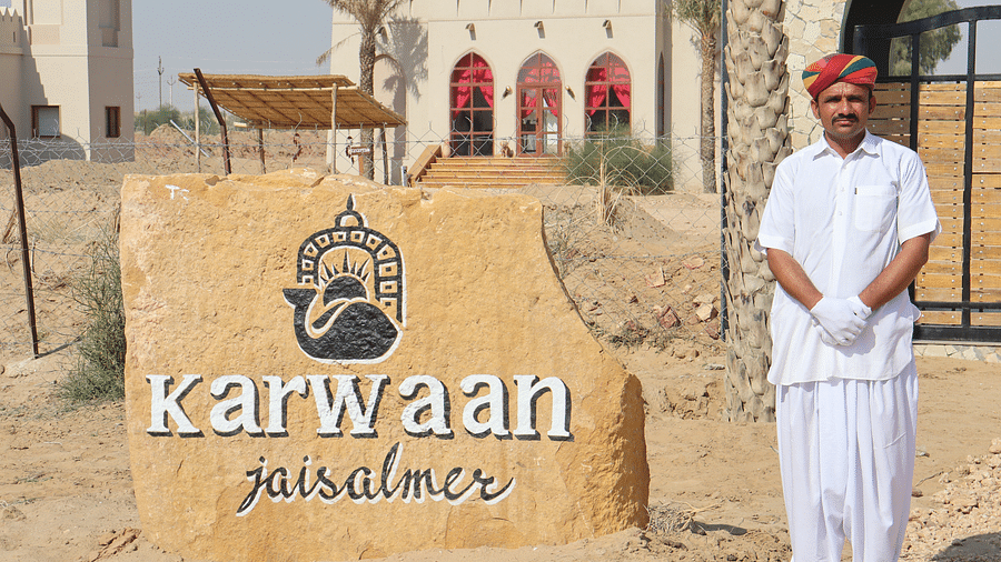 A hotel staff dressed in ethnic wear stands next to the Karwaan Jaisalmer logo printed on a rock 2