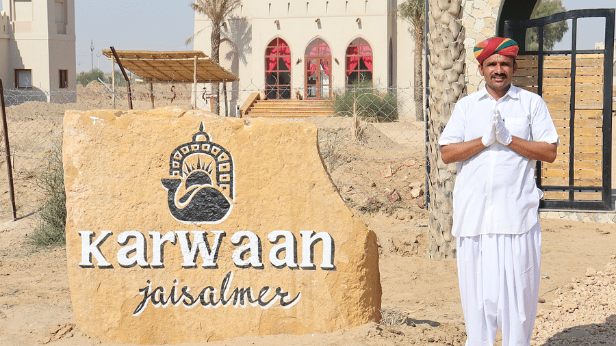 A hotel staff dressed in ethnic wear stands next to the Karwaan Jaisalmer logo printed on a rock 2 