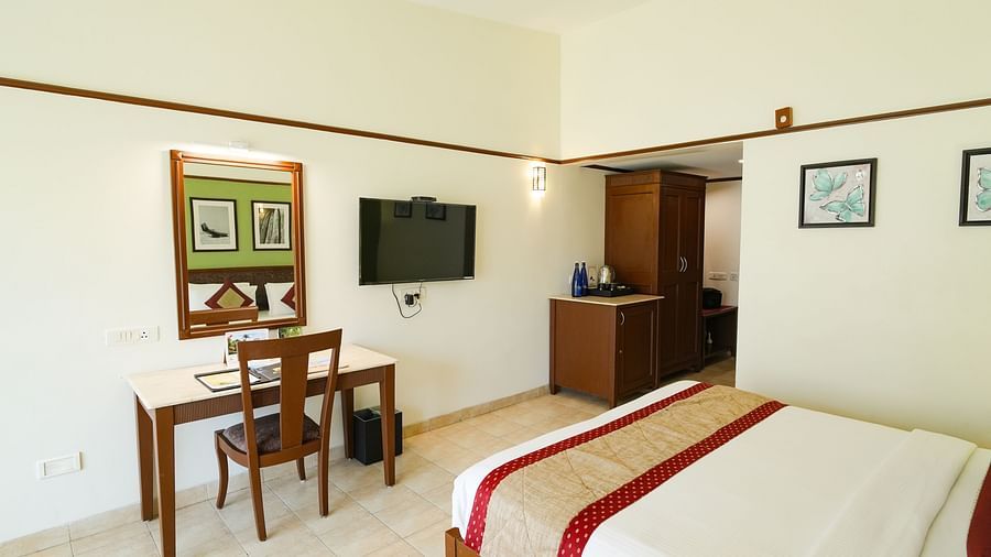 A bright bedroom featuring a bed, a study table with a chair and a mirror in front, a wall-mounted TV, and a wardrobe