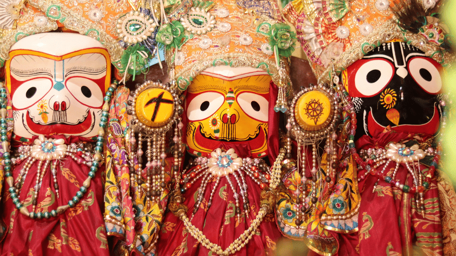 Three colourful, traditional wooden idols of Jagannath, Balabhadra, and Subhadra from the Hindu tradition, adorned with elaborate crowns, jewellery, and red patterned silk garments.