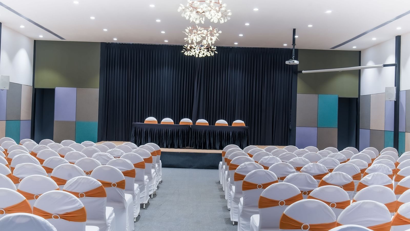 Royal Affair at Darza Resort features numerous white chairs indoor with orange sashes facing a stage with a draped backdrop and several chairs.