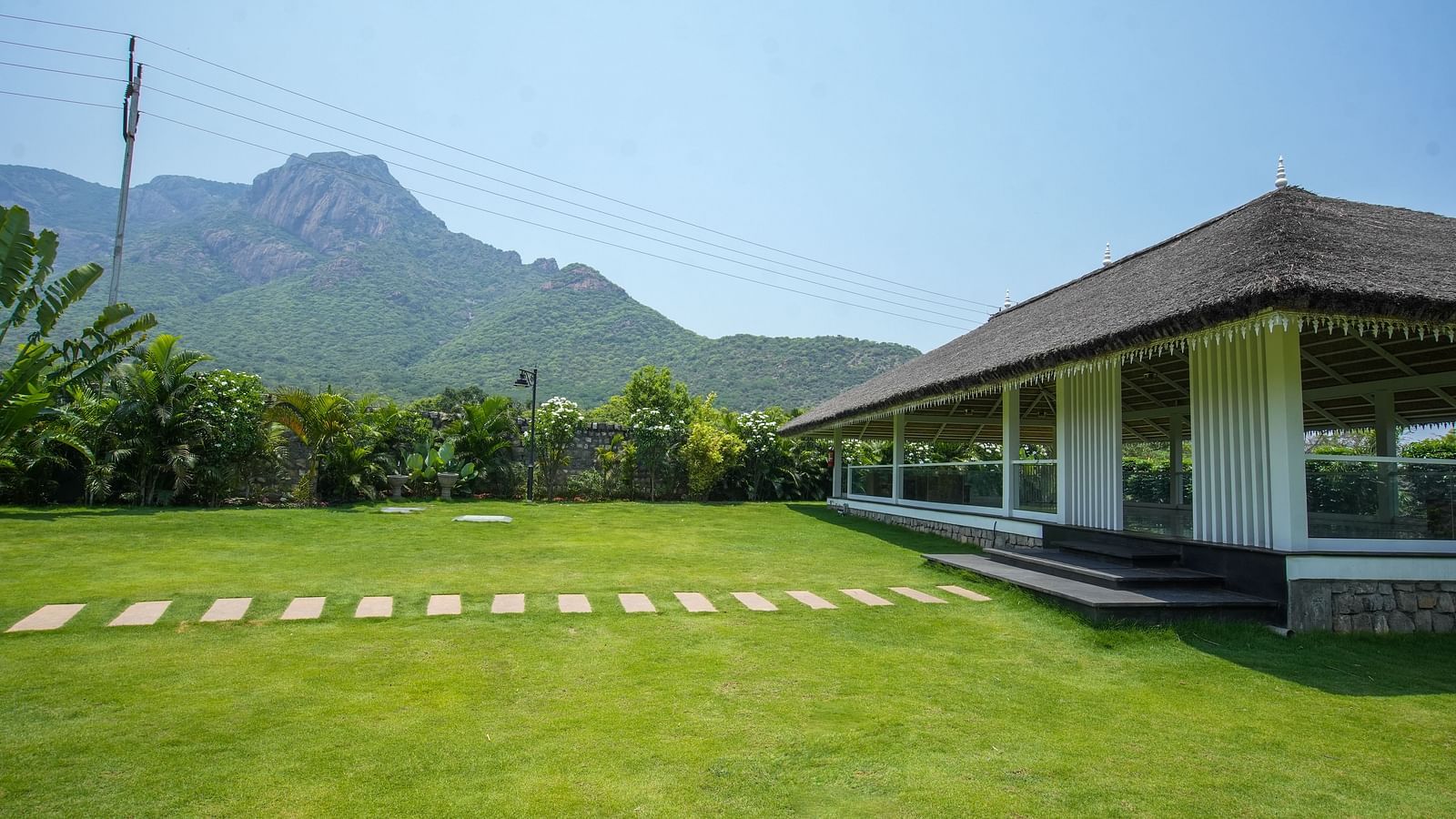 Slice of Heaven at Darza Resort featuring a bright green lawn with a stone pathway leads to a white building with a thatched roof, set against a backdrop of lush greenery and mountains.