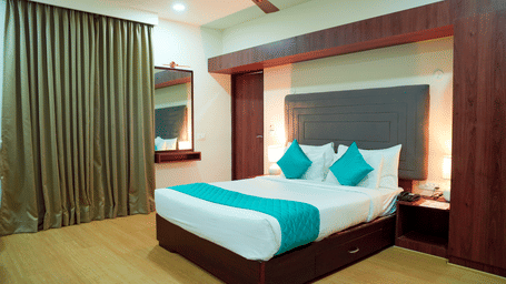 An indoor view shows a bed, pillows, a ceiling fan, curtains, and bedside tables at DNC Shevaroys Resorts and Spa.