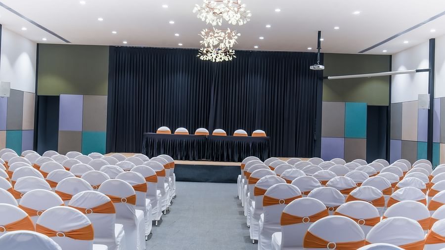Royal Affair at Darza Resort features numerous white chairs indoor with orange sashes facing a stage with a draped backdrop and several chairs.