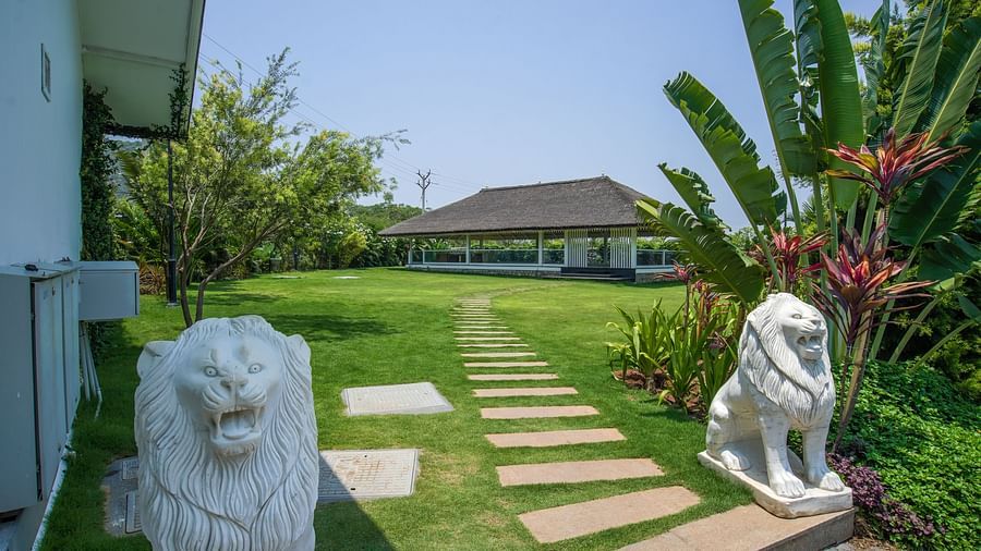 Slice of Heaven at Darza Resort featuring a stone pathway, flanked by two white lion statues, leads across a green lawn towards an open-sided building amidst lush tropical foliage under a bright sky.