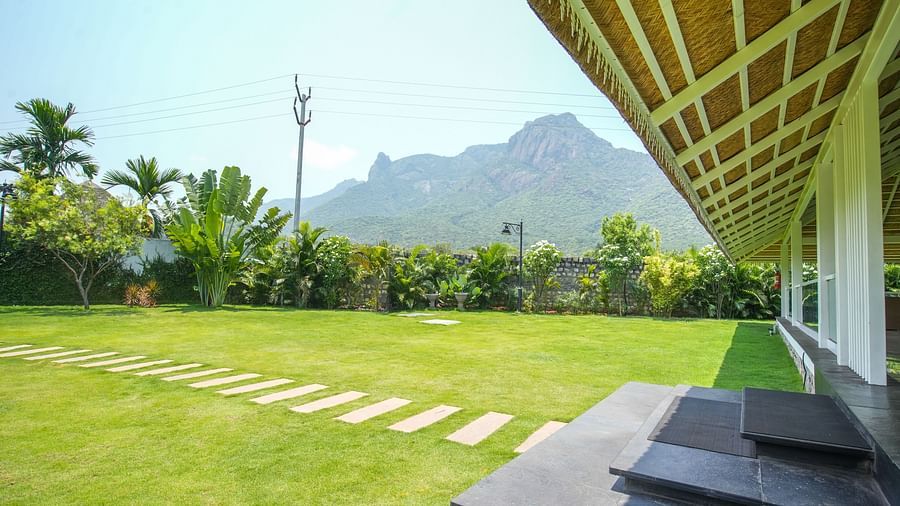 Slice of Heaven at Darza Resort featuring a covered porch with a bamboo roof, from where a stone pathway leads across a green lawn towards lush vegetation and a large mountain under a clear sky.