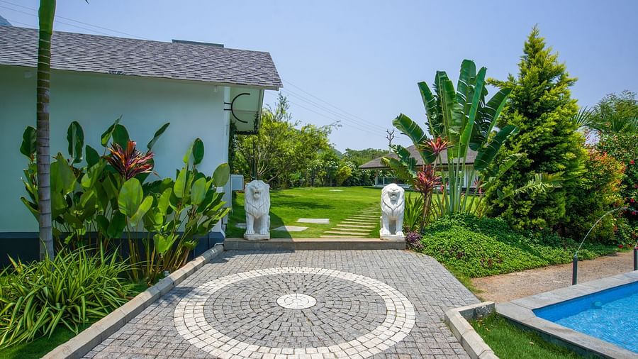 Slice of Heaven at Darza Resort featuring stone pathway leading past two white elephant statues towards a green lawn and lush tropical plants under a bright sky, with a glimpse of a blue pool.