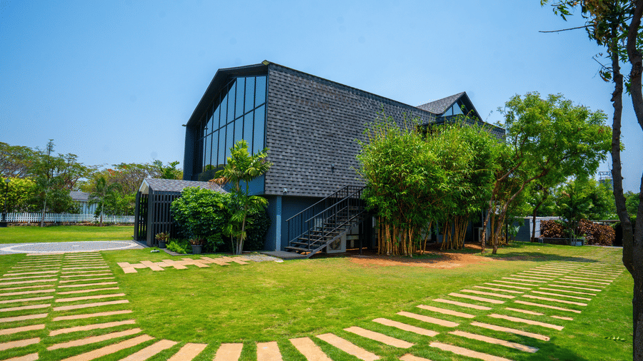 Facade of Darza Resort with large windows is nestled amongst bright green lawns and trees under a clear, sunny sky, with stone pathways leading across the grass.