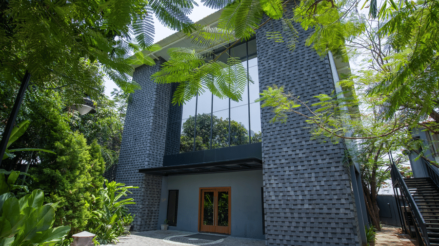 Facade of Royal Affair at Darza Resort with textured grey walls and large glass windows is partially obscured by lush green foliage under a bright sky. - Business Resort Coimbatore
