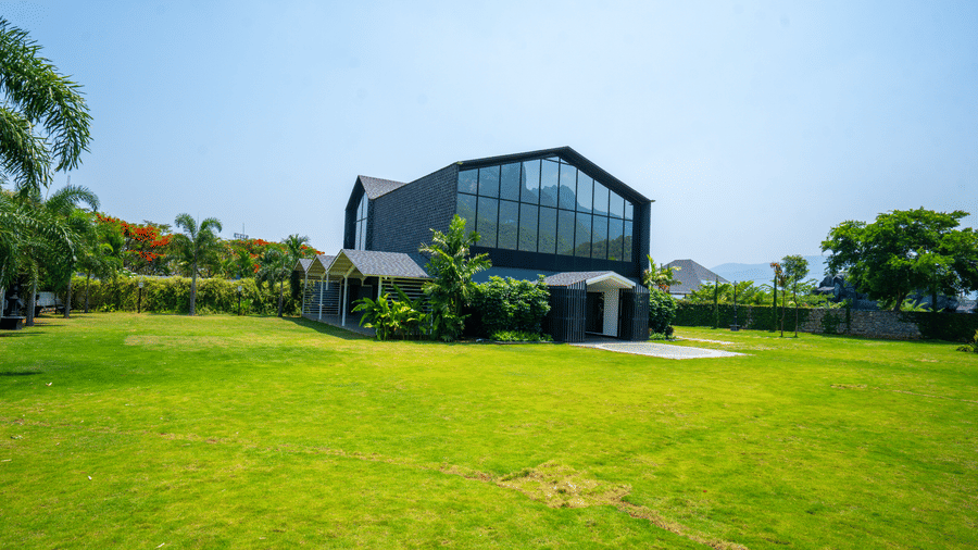 Facade of Love Letter lawn at Darza Resort with a building in the centre having large glass panes standing on a sprawling green lawn under a bright blue sky, surrounded by greenery.