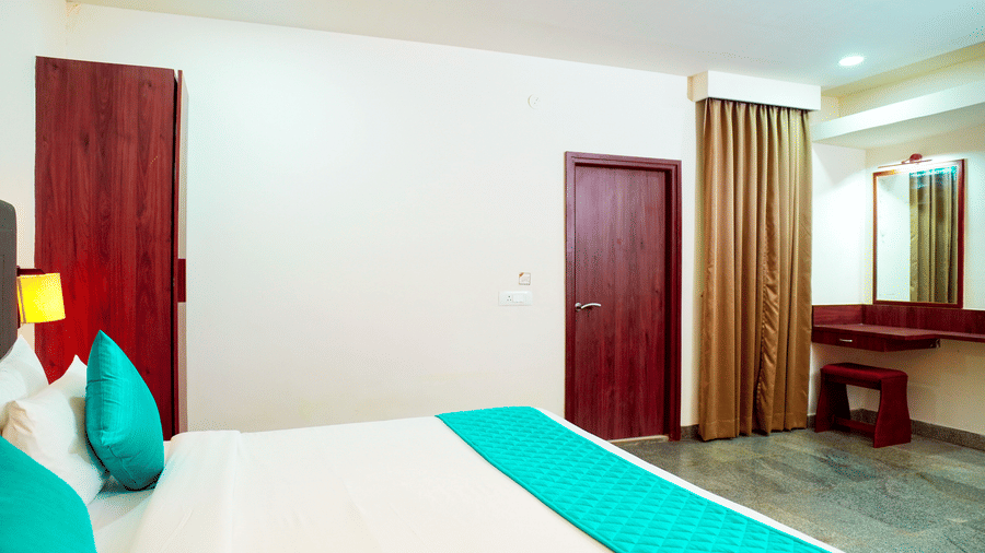 An indoor view shows a bed, pillows, a door, a curtain, and a dressing area at DNC Shevaroys Resorts and Spa.