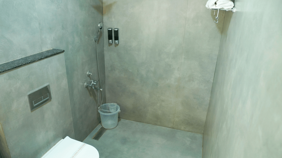 A bathroom view shows a toilet, a shower area, a bucket, and a towel on a holder at DNC Shevaroys Resorts and Spa.