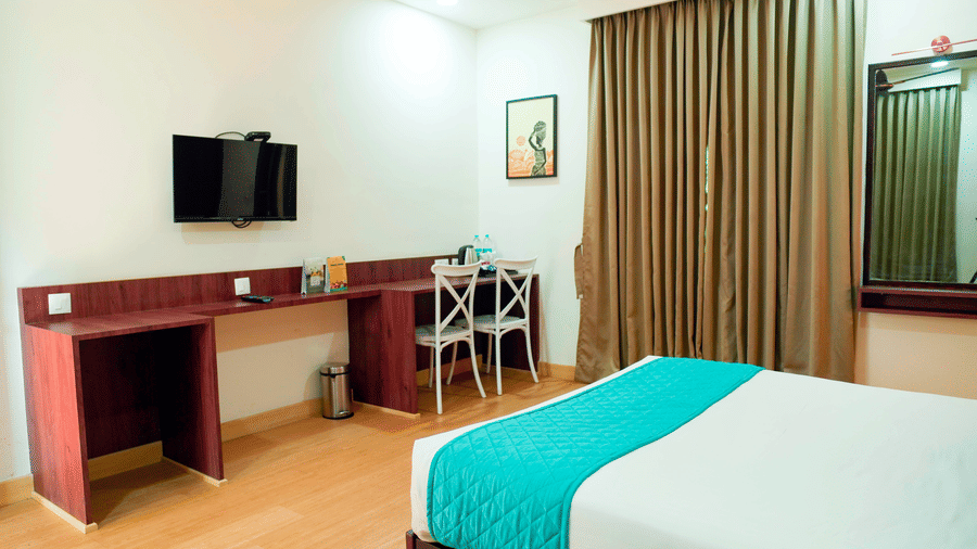 An indoor view shows a bed, a desk with a television, a chair, curtains, and wall art at DNC Shevaroys Resorts and Spa.