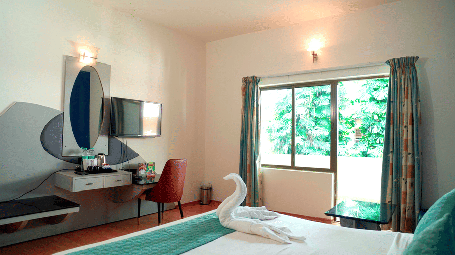 An indoor view shows a bed, a towel arrangement, a dressing table, a mirror, and a window at DNC Shevaroys Resorts and Spa.