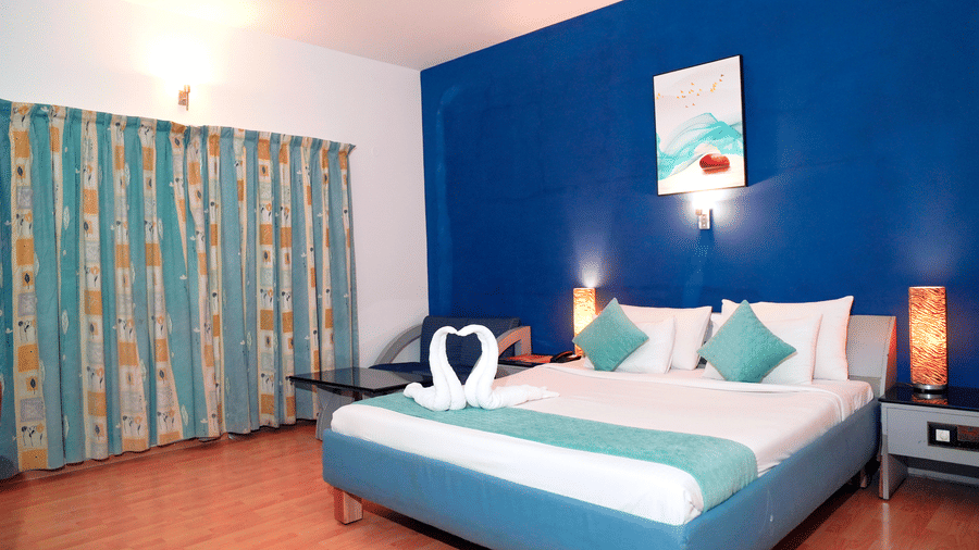 An indoor view shows a bed, pillows, a wall painting, a ceiling fan, and curtains at DNC Shevaroys Resorts and Spa.