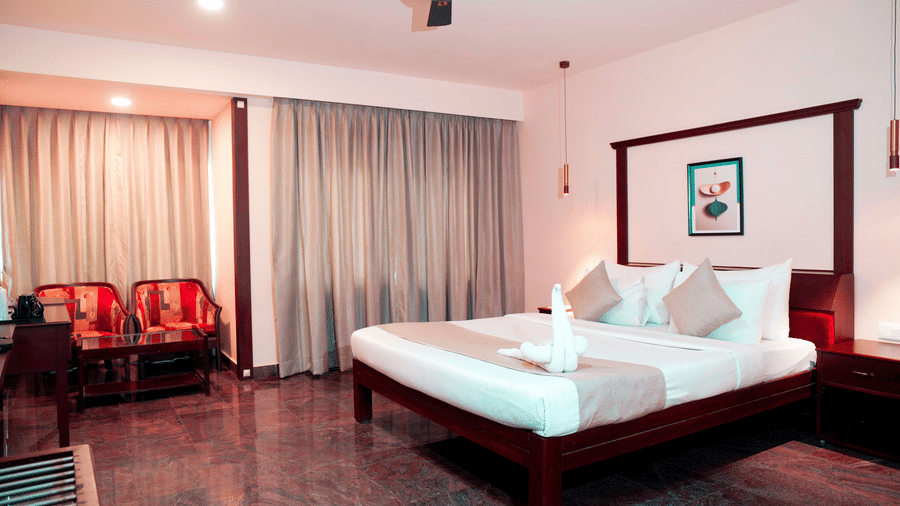 An indoor view shows a bed, a ceiling fan, chairs, a table, and curtains at DNC Shevaroys Resorts and Spa.