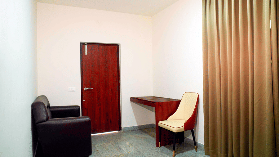 A simple guest room interior at DNC Shevaroys Resort & Spa featuring a dark sofa, a wooden desk, and full-length brown curtains.
