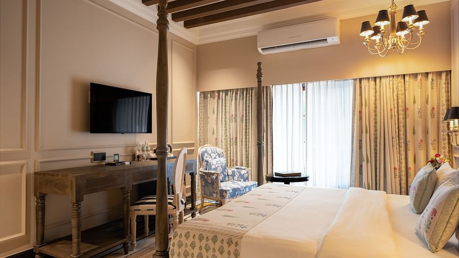 Elegant hotel room with a four-poster bed, floral curtains, a chandelier, and wooden ceiling beams. The room features a wall-mounted TV, a wooden desk with chairs, and a blue floral armchair by large windows with sheer drapes - Panna Vilas Palace