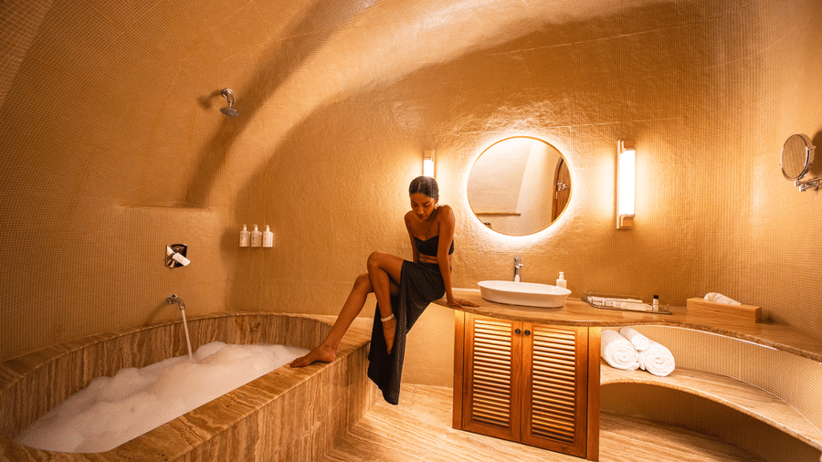 A luxury cave-style bathroom featuring a sunken bathtub filled with foam, a glowing round mirror, and a woman sitting on the vanity counter.