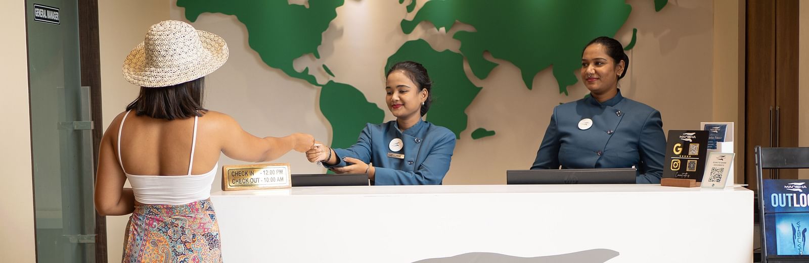 The reception area of a hotel with staff members and guests and a world map on the wall | Mansha Hotels