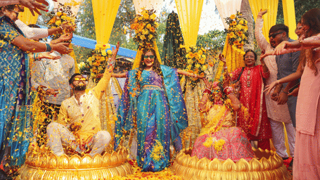 A vibrant wedding ceremony with a couple seated on gold platforms as friends and family shower them with yellow and orange flower petals under a canopy.