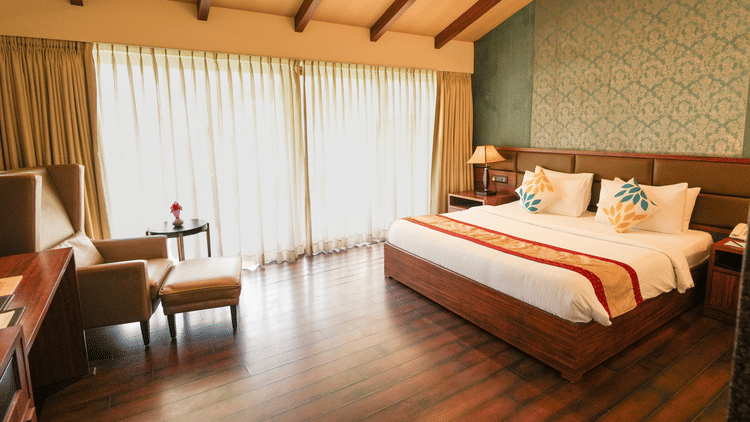 A hotel room with a neatly arranged double bed, a bedside table with a lamp, long curtains, aesthetic interiors, a recliner sofa, a coffee table with a flower vase, and a study table on the side