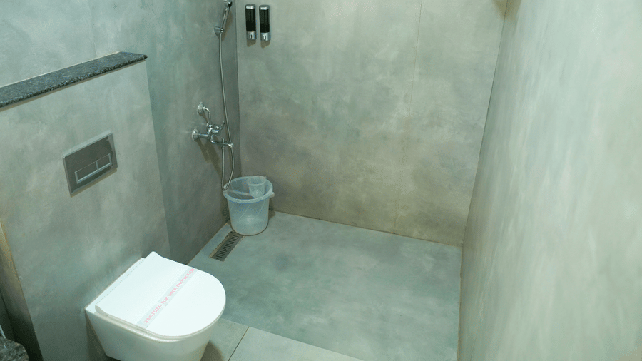 A bathroom view shows a toilet, a shower area, a bucket, and a towel on a holder at DNC Shevaroys Resorts and Spa.