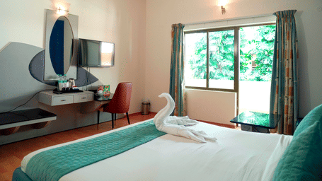 An indoor view shows a bed, a towel arrangement, a dressing table, a mirror, and a window at DNC Shevaroys Resorts and Spa.